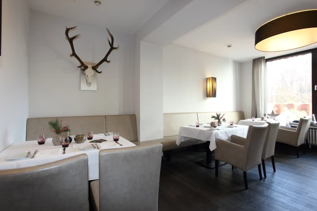 Heller Gastraum mit weiß eingedeckten Tischen, beigen Polstermöbeln und Hirschgeweih an der Wand.