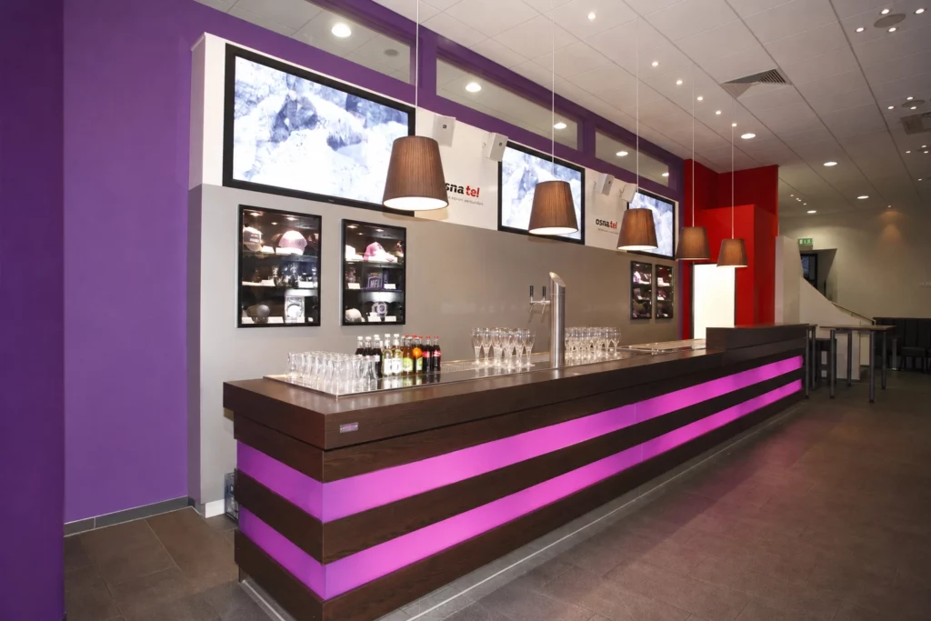 Lange Holztheke mit pinkfarbenen Lichtbändern, Zapfsäule und Gläsern vor violetter Wand.